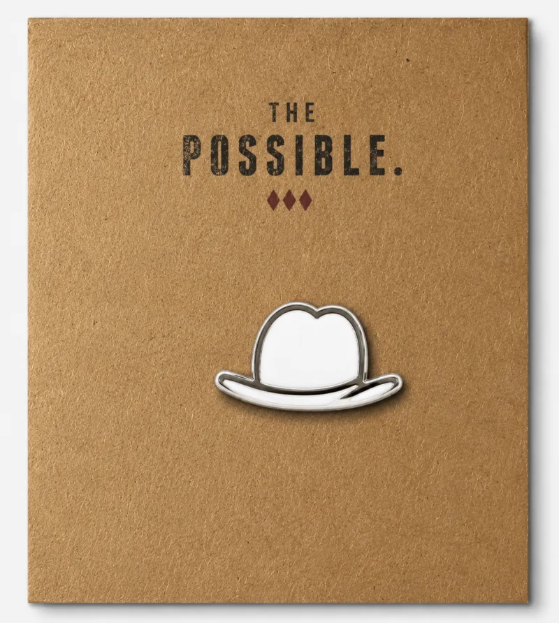 The Possible enamel lapel pin — the white fedora in miniature, on a kraft card.
